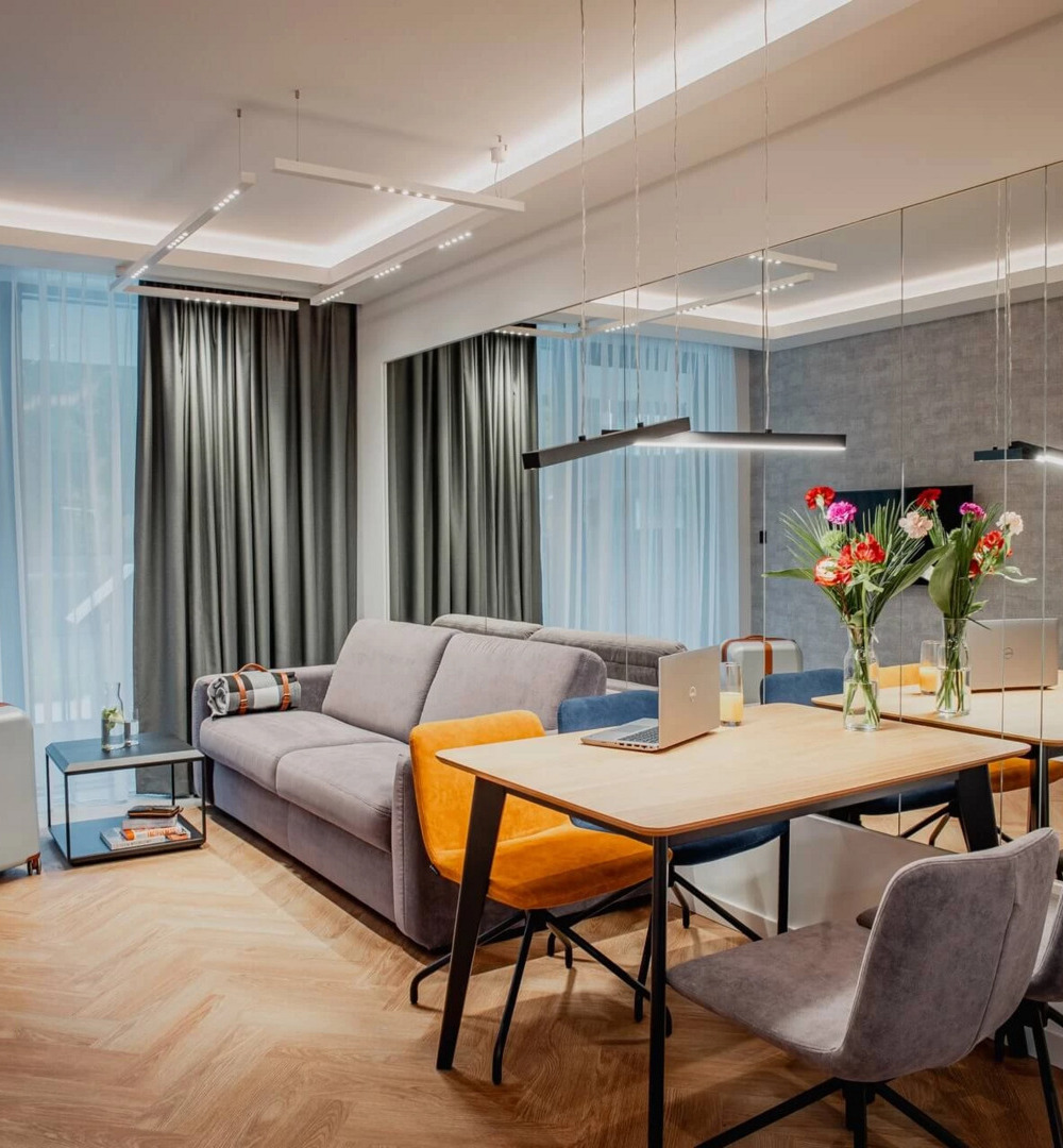 Nowoczesny apartament z szarą sofą, kolorowymi krzesłami przy stole, laptopem i bukietem kwiatów, jasne zasłony i drewniana podłoga