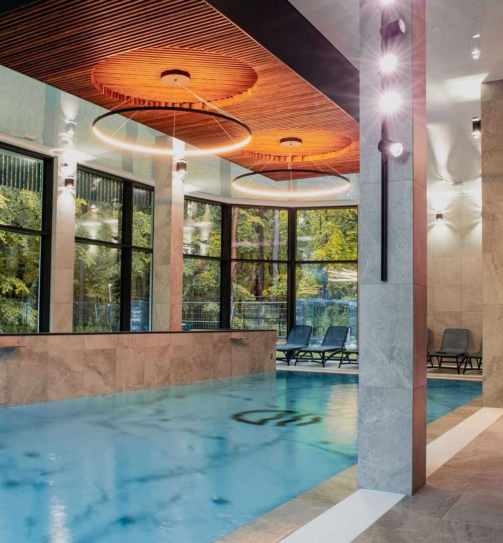Nowoczesny basen wewnętrzny z dużymi oknami na zielony ogród, stylowymi krzesłami do relaksu i drewnianym sufitem z oświetleniem LED