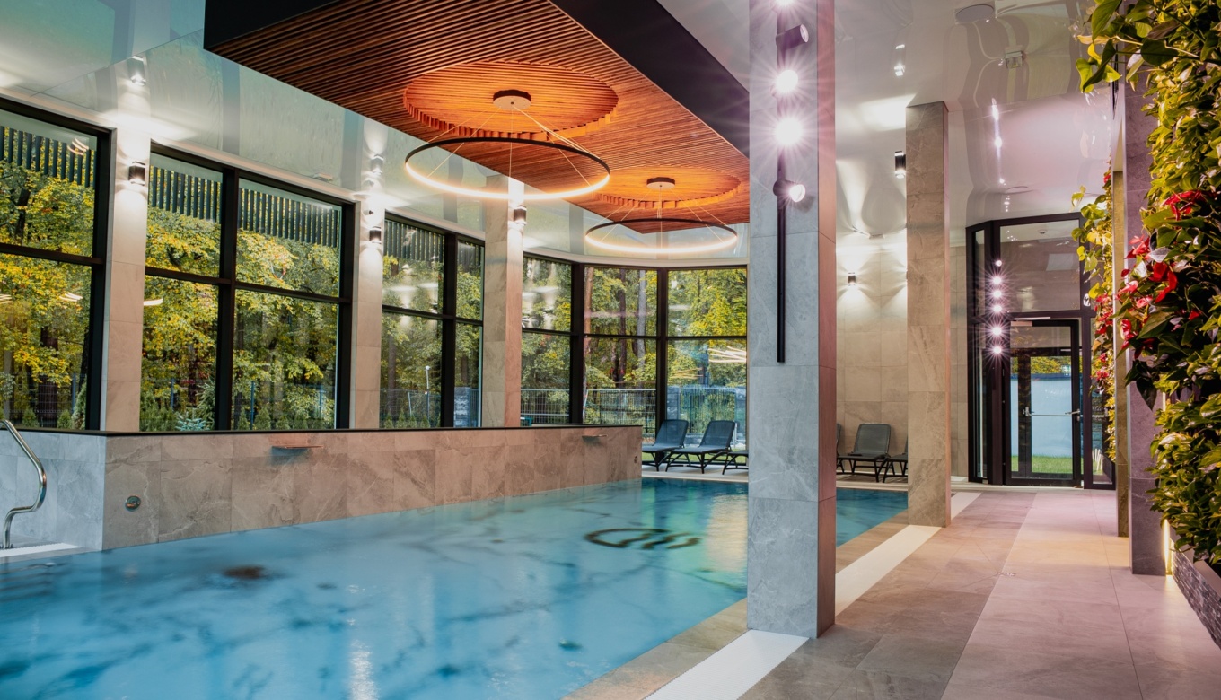 Nowoczesny basen wewnętrzny z dużymi oknami na zielony ogród, stylowymi krzesłami do relaksu i drewnianym sufitem z oświetleniem LED
