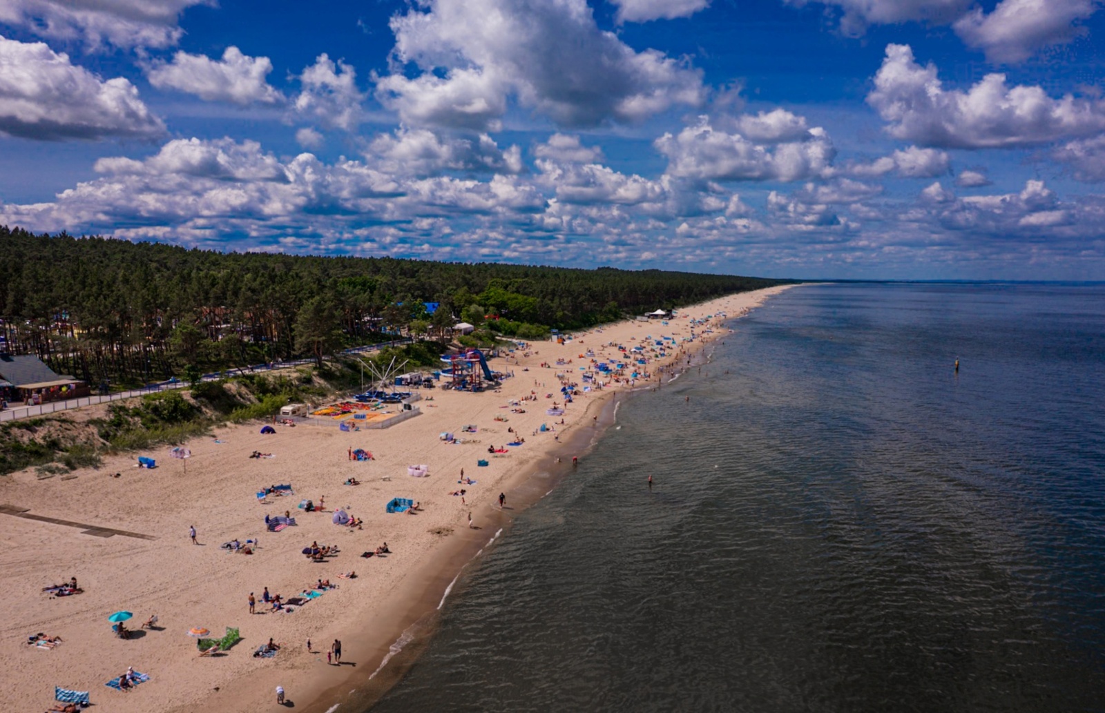 Długi piaszczysty plaża z gośćmi plażującymi i lasem w tle, idealna na relaksujący wypoczynek nad morzem