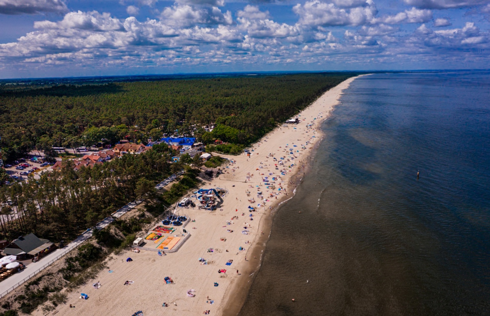 Długa piaszczysta plaża z rozproszonymi parawanami i gośćmi, obok las sosnowy i małe budynki wypoczynkowe