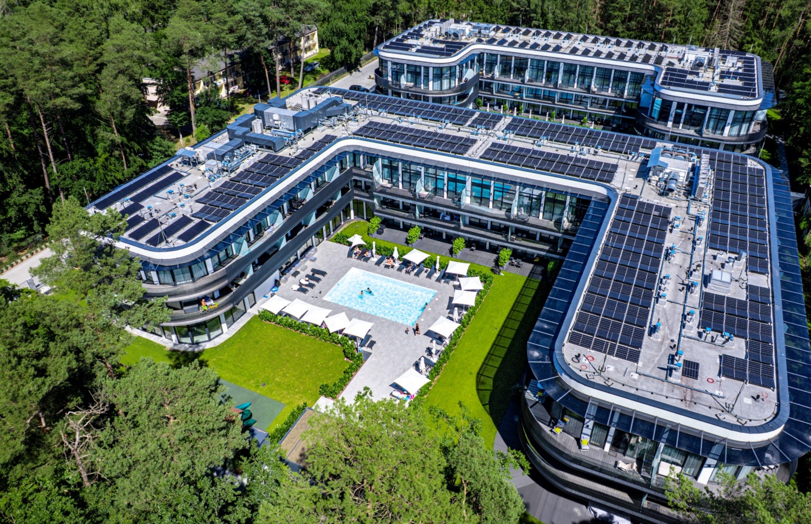 Nowoczesny hotel z dużym basenem na dziedzińcu, otoczony lasem, z tarasami i panelami słonecznymi na dachu
