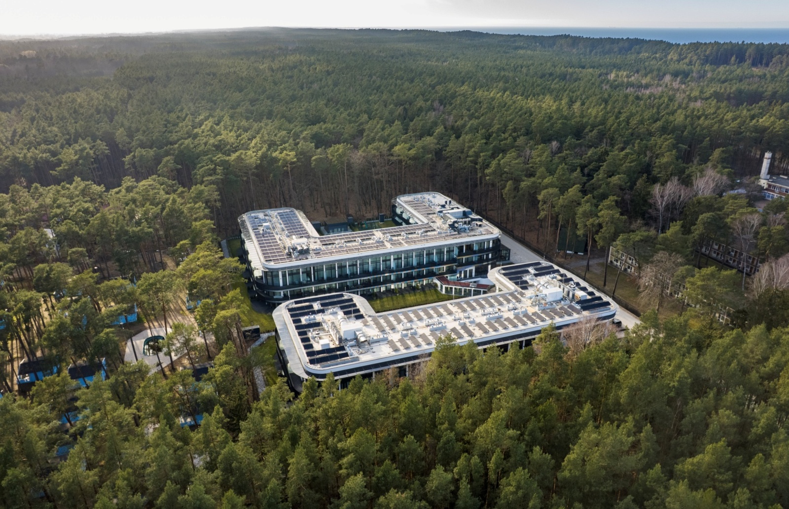 Nowoczesny hotel z przeszklonymi fasadami otoczony gęstym lasem sosnowym, z widokiem na morze w tle