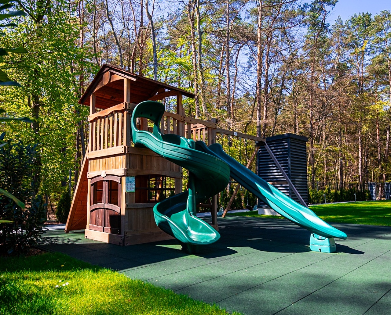 Plac zabaw z drewnianą wieżą i dwoma zielonymi zjeżdżalniami na tle lasu, idealny dla rodzin z dziećmi