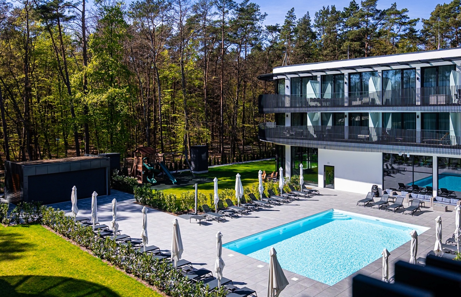 Nowoczesny hotel z odkrytym basenem, leżakami i parasolami na tarasie, otoczony lasem i zielonym trawnikiem.