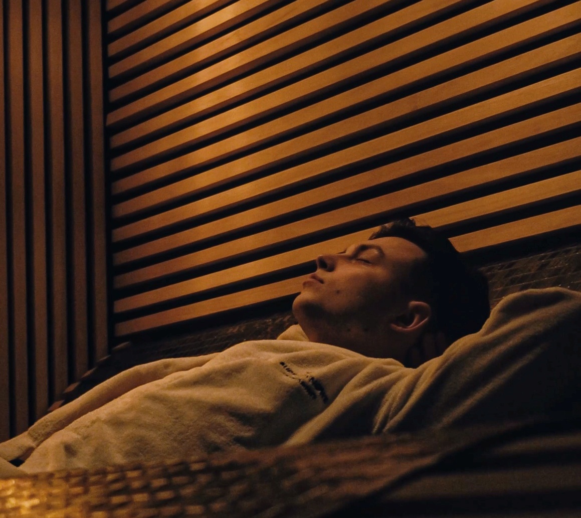 Mężczyzna w białym szlafroku wypoczywa na leżance w ciepłym, drewnianym wnętrzu spa, emanacja relaksu.