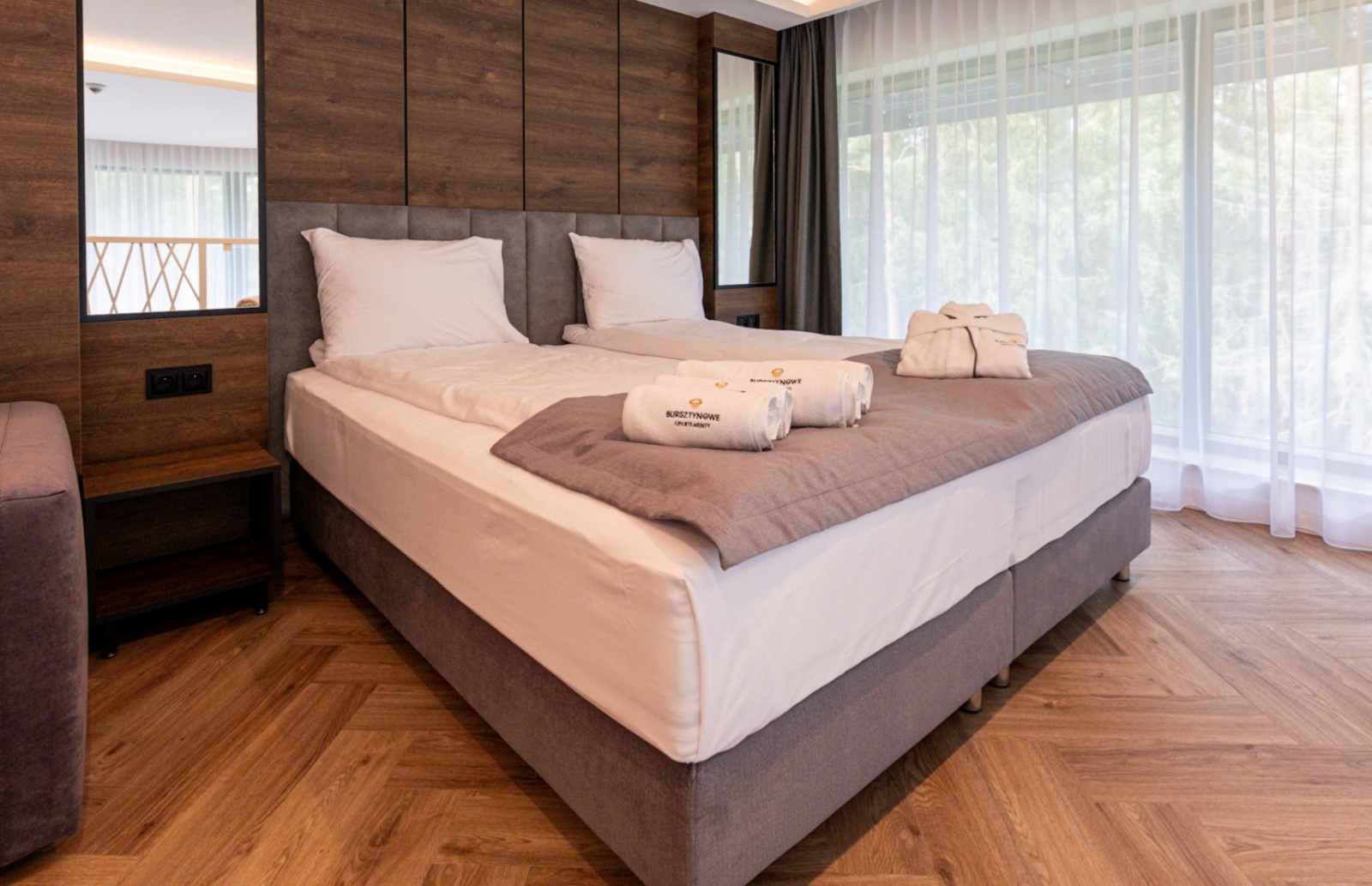 Przytulny pokój z dużym łóżkiem, ręcznikami i szlafrokami, drewniana podłoga i okno z widokiem na zieleń.