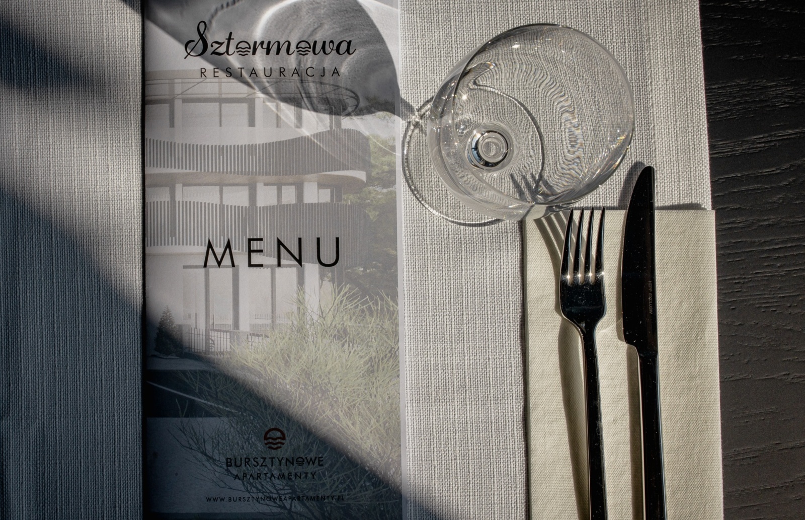 Elegancko nakryty stół z menu restauracji Szterma i sztućcami na jasnym obrusie, szkło odbija światło