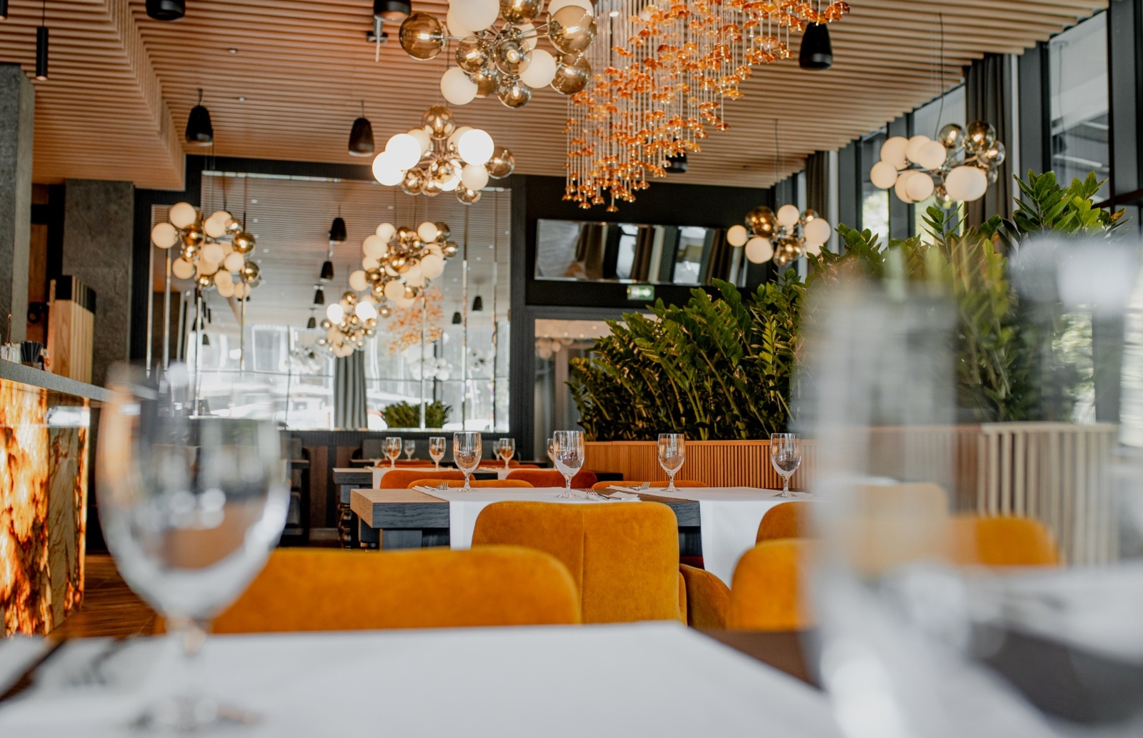 Nowoczesna restauracja z pomarańczowymi krzesłami, białymi obrusami, efektownymi żyrandolami i zielonymi roślinami we wnętrzu