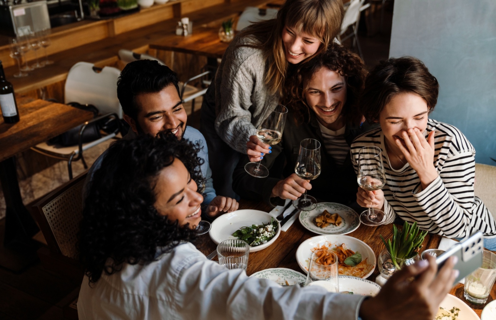 Grupa przyjaciół w przytulnej restauracji robiąca selfie przy stole z przekąskami i kieliszkami z winem.
