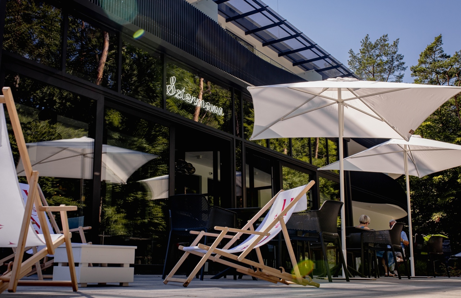 Taras hotelowy z drewnianymi leżakami, białymi parasolami i stolikami na zewnątrz w otoczeniu zielonych drzew