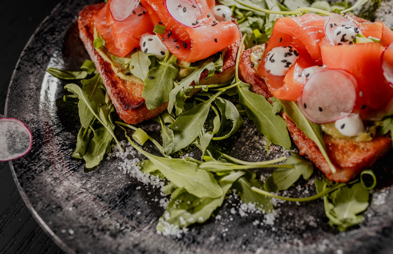 Tosty z wędzonym łososiem, rukolą, kremowym sosem i cienkimi plasterkami rzodkiewki na ciemnym talerzu