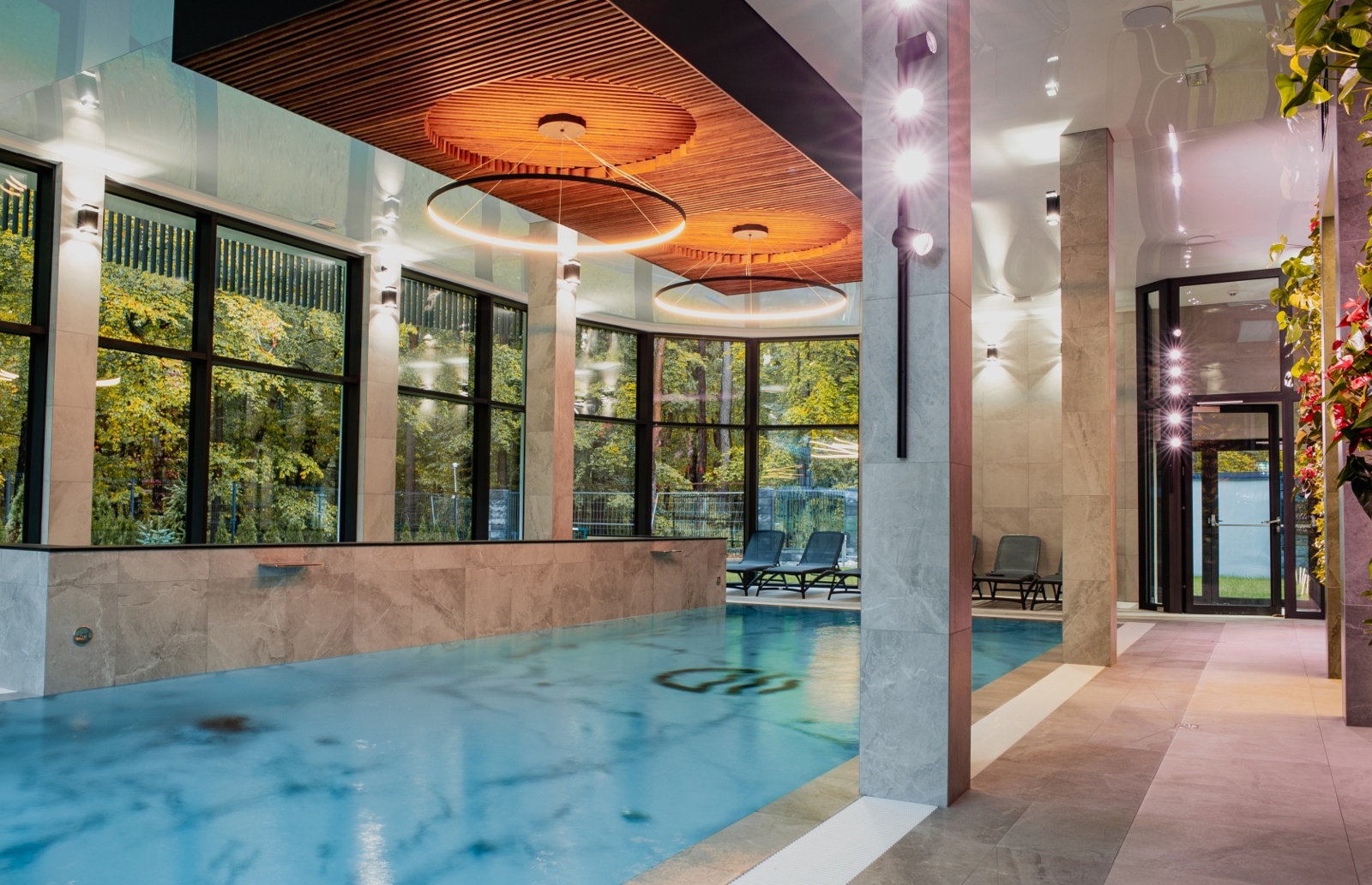 Nowoczesny basen wewnętrzny z dużymi oknami na zielony ogród, stylowymi krzesłami do relaksu i drewnianym sufitem z oświetleniem LED