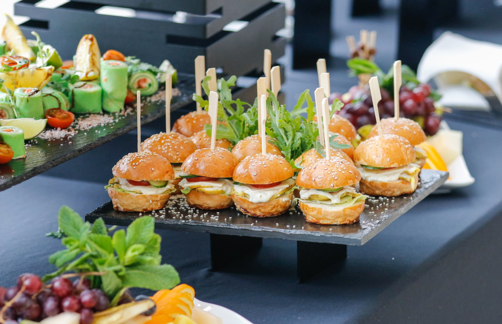 Mini burgers z sezamowymi bułkami, rukolą i warzywami na eleganckiej tacy, obok przekąski i świeże owoce na bufecie