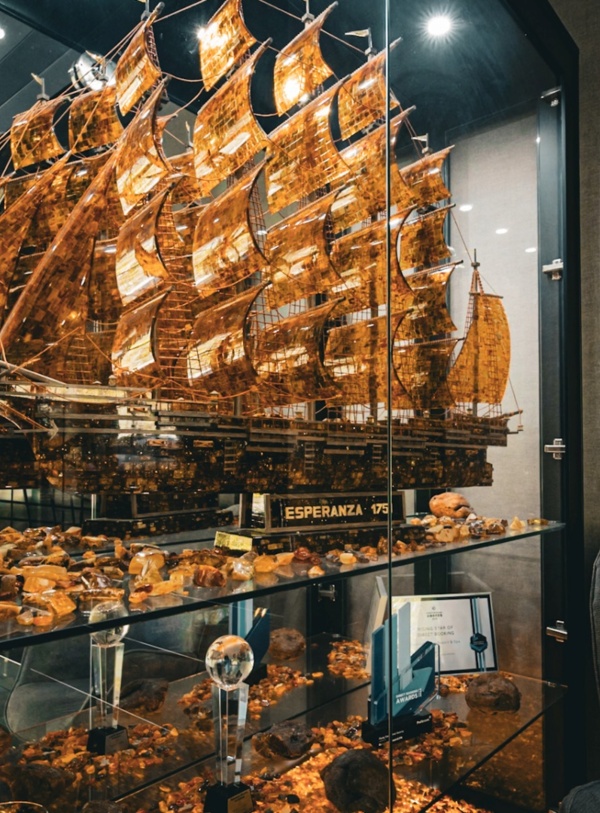 Szczegółowy model statku Esperanza 175 z bursztynowymi żaglami w eleganckiej gablocie hotelowego lobby z wygodnym fotelem obok.