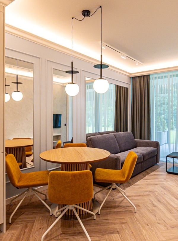 Nowoczesny apartament z aneksem kuchennym, okrągłym stołem, czterema żółtymi fotelami i szarą sofą przy dużym oknie z widokiem na zieleń.