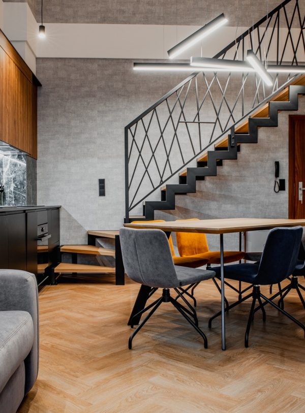 Nowoczesny apartament z aneksem kuchennym, jadalnią z kolorowymi krzesłami, szarą sofą i modnym oświetleniem pod schodami