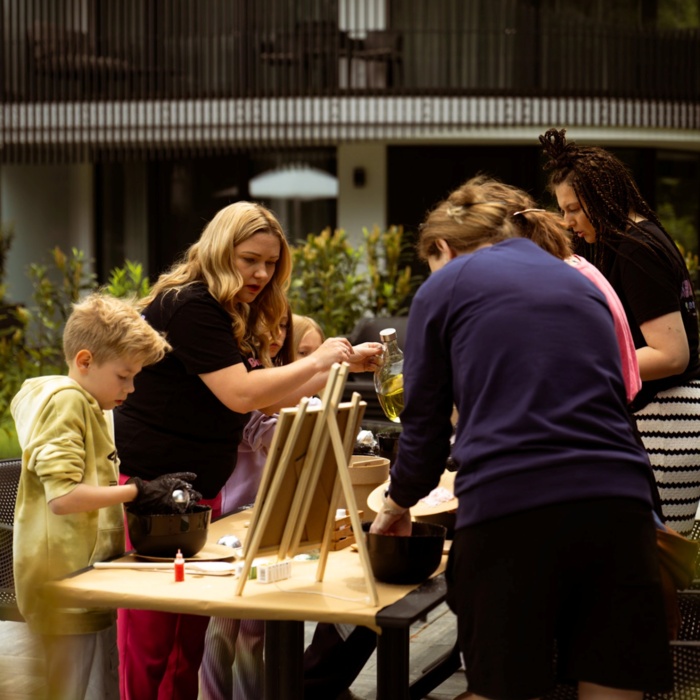 Grupa gości uczestniczy w warsztatach kulinarnych na zewnątrz, przy stole z akcesoriami do gotowania, na tarasie hotelowym.