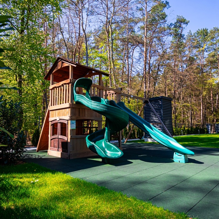 Plac zabaw z drewnianą wieżą i dwoma zielonymi zjeżdżalniami na tle lasu, idealny dla rodzin z dziećmi