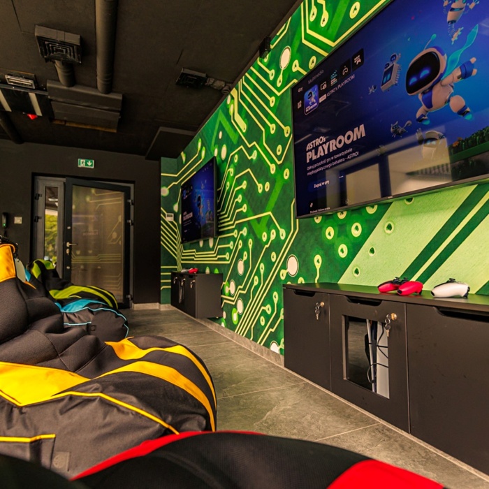 Nowoczesny pokój rozrywki z kolorowymi pufami, dwoma telewizorami na ścianie z motywem obwodów elektronicznych oraz konsolami do gier.