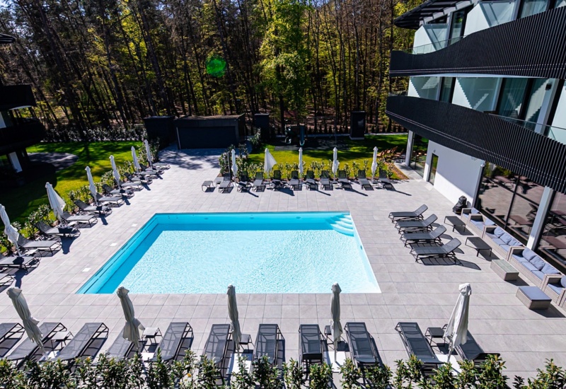 Nowoczesny hotel z basenem na dziedzińcu, otoczony leżakami i parasolami, w otoczeniu zieleni i lasu