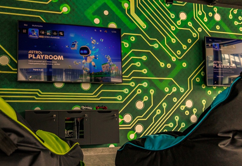 Nowoczesny pokój gier z zieloną ścianą ozdobioną wzorem obwodów elektronicznych i wygodnymi fotelami typu pufy.