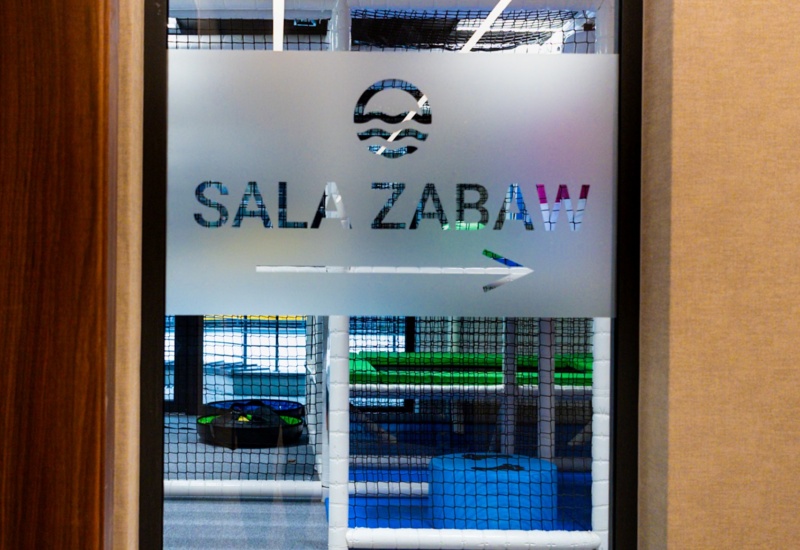 Tablica wskazuje drogę do kolorowej, bezpiecznej sali zabaw dla dzieci z miękkimi siedziskami i siatką ochronną