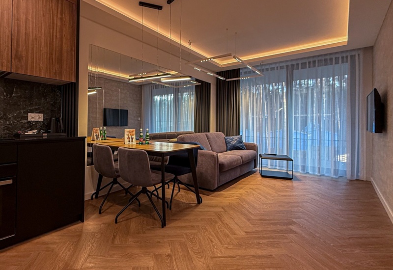 Nowoczesny pokój dzienny z aneksem kuchennym, miękką sofą, stołem jadalnianym na cztery osoby i dużymi oknami z zasłonami.