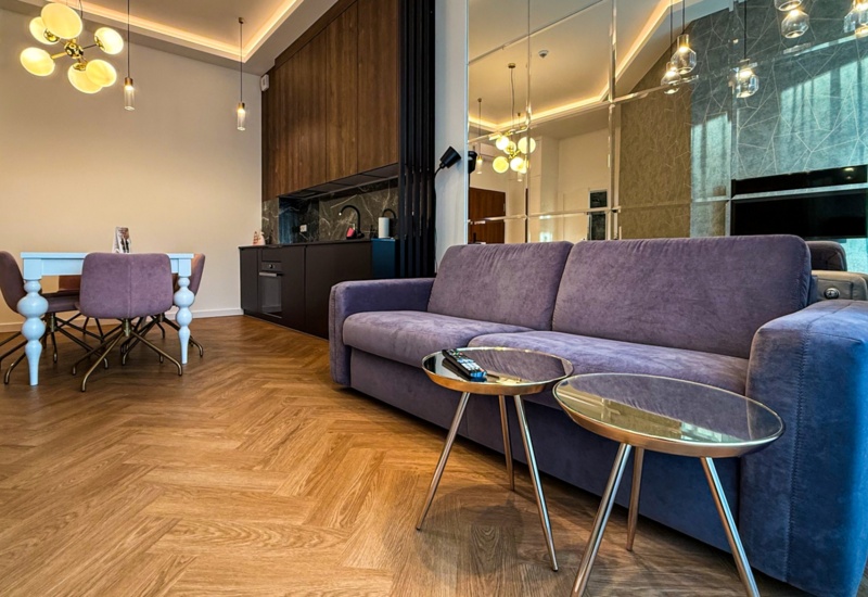 Nowoczesny pokój hotelowy z fioletową sofą, stolikami, białym stołem i fioletowymi krzesłami, kuchnia z drewnianymi szafkami