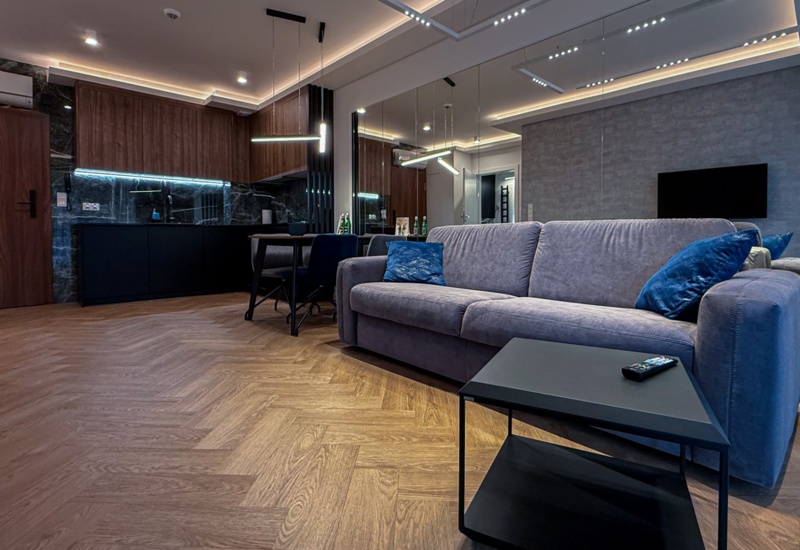 Nowoczesny apartament z salonem, szarą sofą, niebieskimi poduszkami, czarnym stolikiem i aneksem kuchennym z marmurowymi płytkami