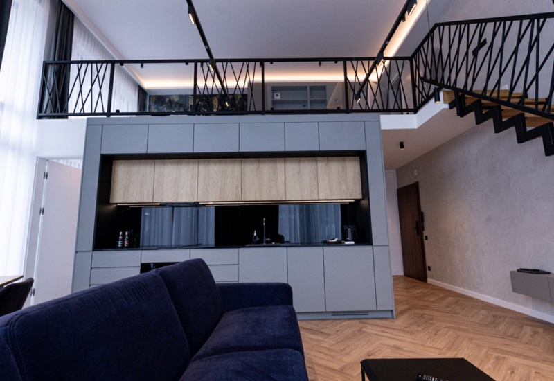 Nowoczesny loft z otwartą kuchnią, szarymi szafkami, granatową sofą i stylową metalową balustradą na antresoli.