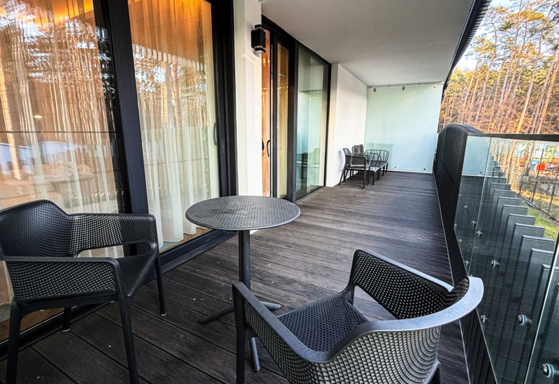 Przestronny balkon hotelowy z czarnymi krzesłami i stolikami, widok na ogród i okoliczny las przez szklaną balustradę