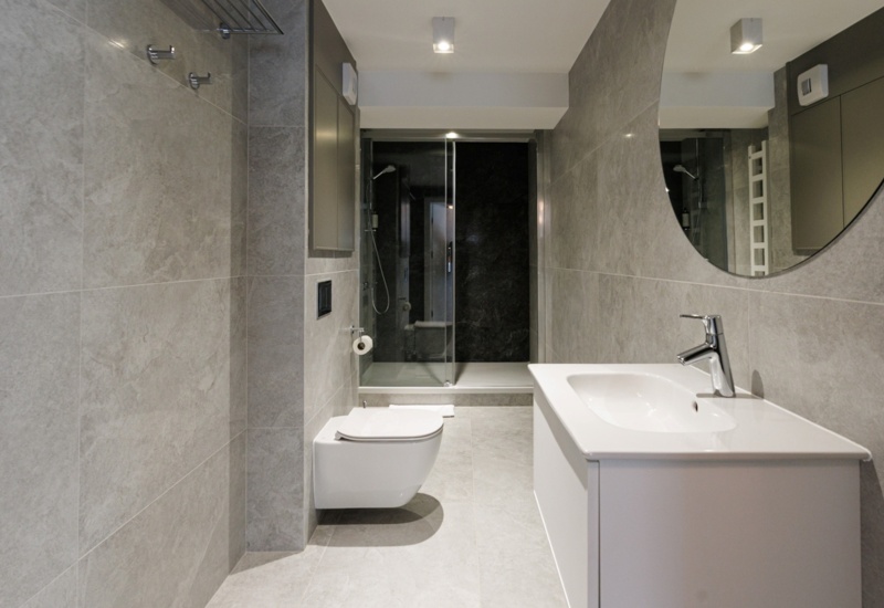 Nowoczesna łazienka hotelowa z szarą glazurą, wiszącą toaletą, dużym lustrem i prysznicem z przeszklonymi drzwiami