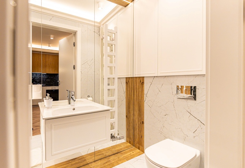 Nowoczesna łazienka hotelowa z białą umywalką, toaletą, drewnianymi akcentami i subtelnym oświetleniem, widok na kuchnię