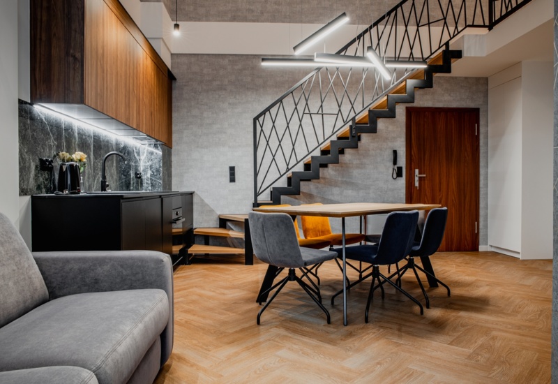 Nowoczesny apartament z aneksem kuchennym, jadalnią z kolorowymi krzesłami, szarą sofą i modnym oświetleniem pod schodami