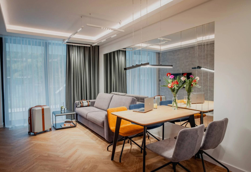 Nowoczesny apartament z szarą sofą, kolorowymi krzesłami przy stole, laptopem i bukietem kwiatów, jasne zasłony i drewniana podłoga