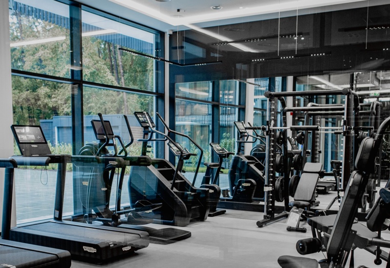 Nowoczesna siłownia hotelowa z bieżniami, rowerkami stacjonarnymi i maszynami do ćwiczeń siłowych przy dużych oknach z widokiem na zieleń.
