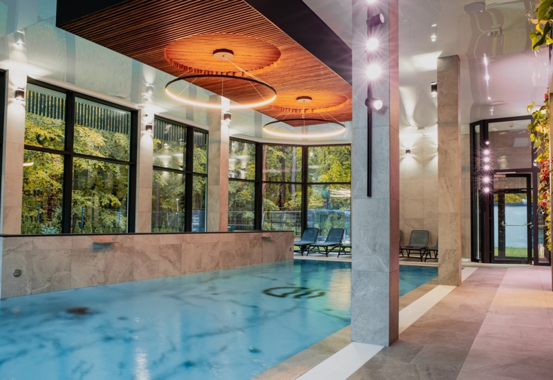 Nowoczesny basen wewnętrzny z dużymi oknami na zielony ogród, stylowymi krzesłami do relaksu i drewnianym sufitem z oświetleniem LED