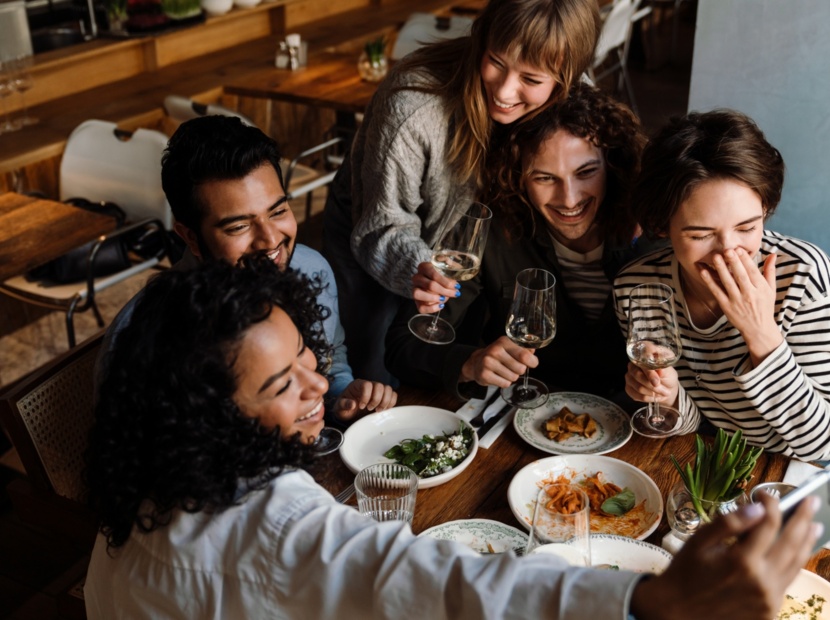 Grupa przyjaciół w przytulnej restauracji robiąca selfie przy stole z przekąskami i kieliszkami z winem.