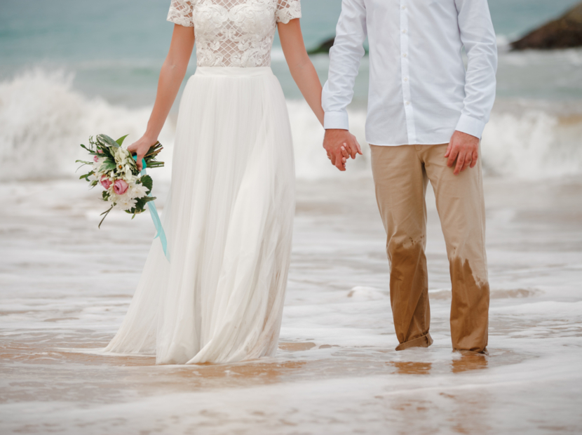 Para młoda trzymająca się za ręce na plaży, panna młoda w białej sukni z bukietem kwiatów, stąpająca po wodzie