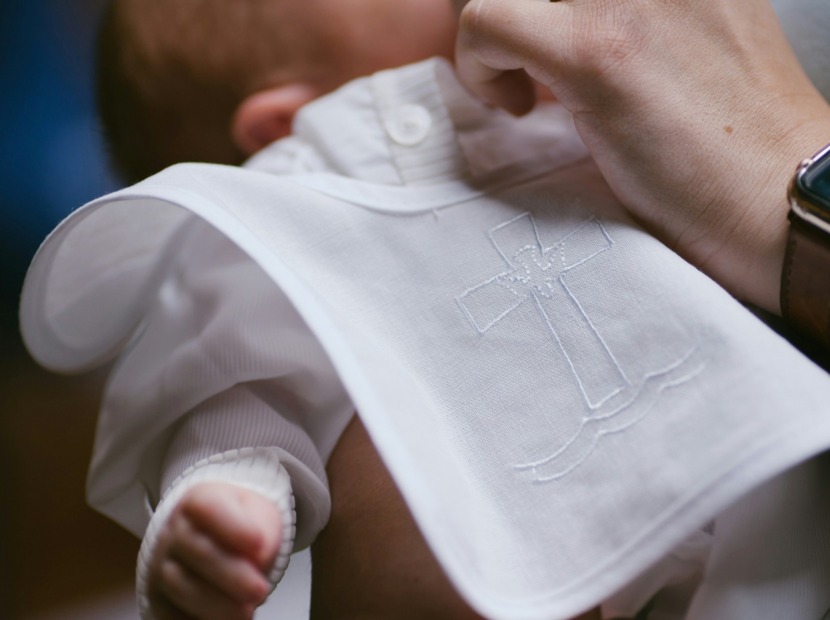 Dziecko ubrane w biały kaftanik z haftowanym krzyżem podczas uroczystości chrztu, w ramionach osoby dorosłej