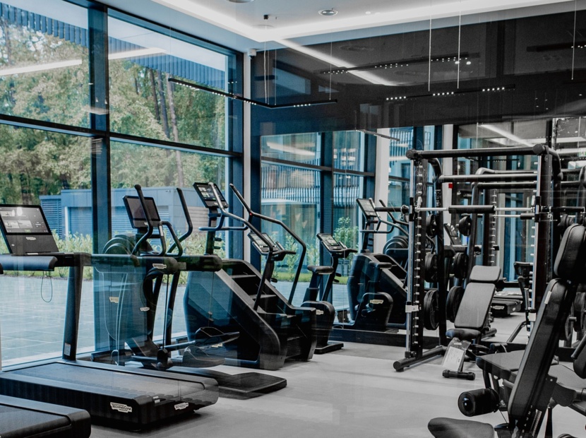 Nowoczesna siłownia hotelowa z bieżniami, rowerkami stacjonarnymi i maszynami do ćwiczeń siłowych przy dużych oknach z widokiem na zieleń.