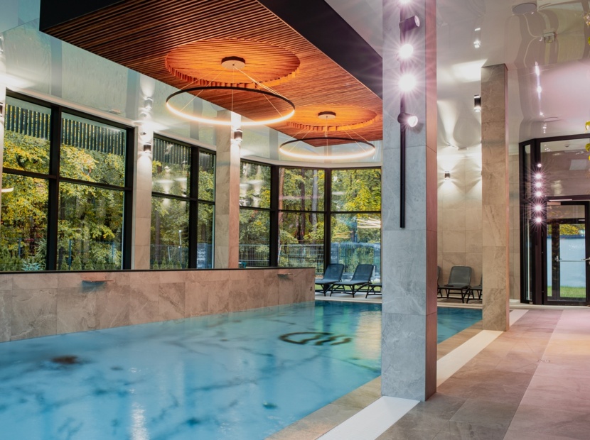 Nowoczesny basen wewnętrzny z dużymi oknami na zielony ogród, stylowymi krzesłami do relaksu i drewnianym sufitem z oświetleniem LED