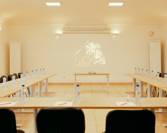 Bright U-shaped meeting room with wooden tables, bottled water and notepads at each seat, projector showing hotel palm logo on wall.
