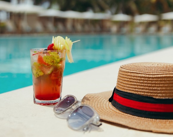 Poolside cocktail with fruit garnish beside a straw sun hat and sunglasses on a resort deck by palm trees