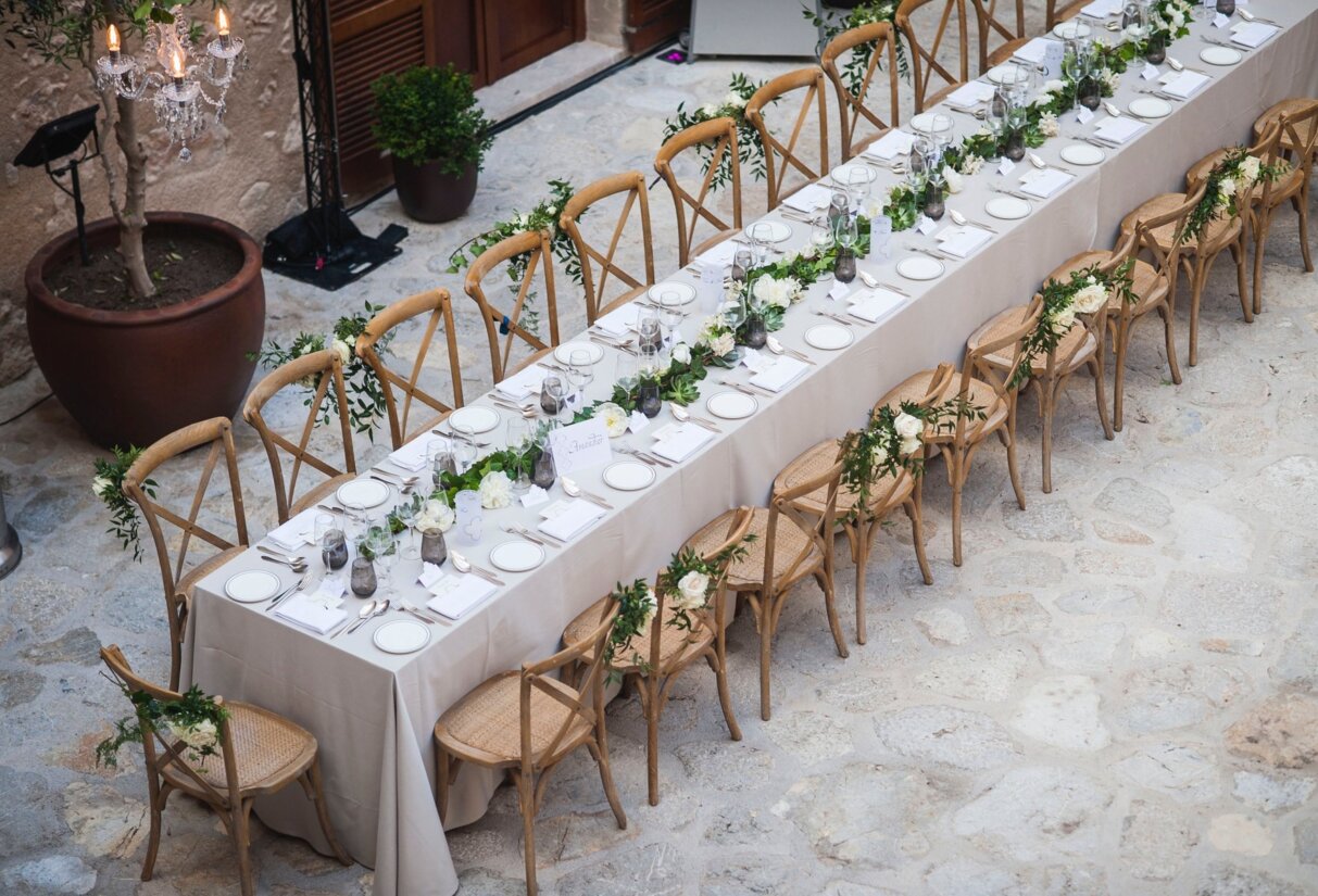 Una larga mesa decorada con plantas y rosas blancas y sillas de madera a los lados
