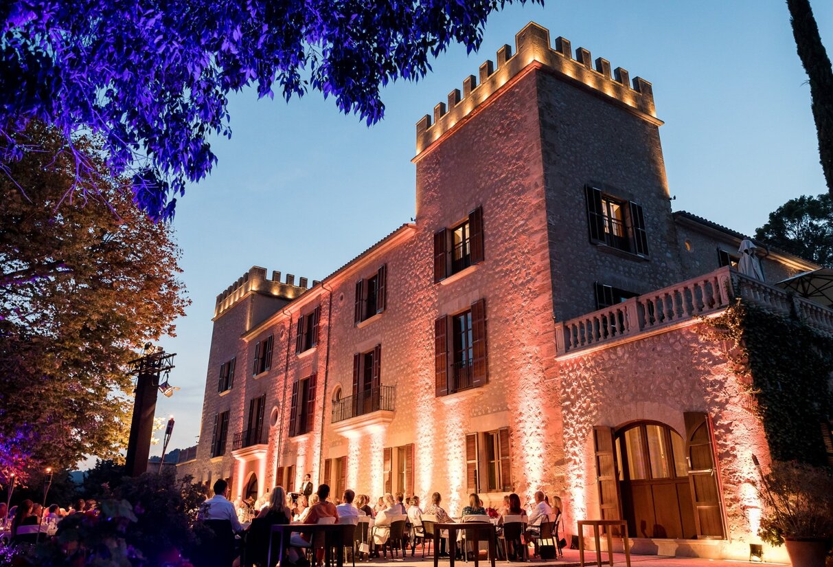 Historisches Steinhotel mit Türmen, abends warm beleuchtet, elegante Freiluftveranstaltung unter leuchtend violetten und orangen Bäumen