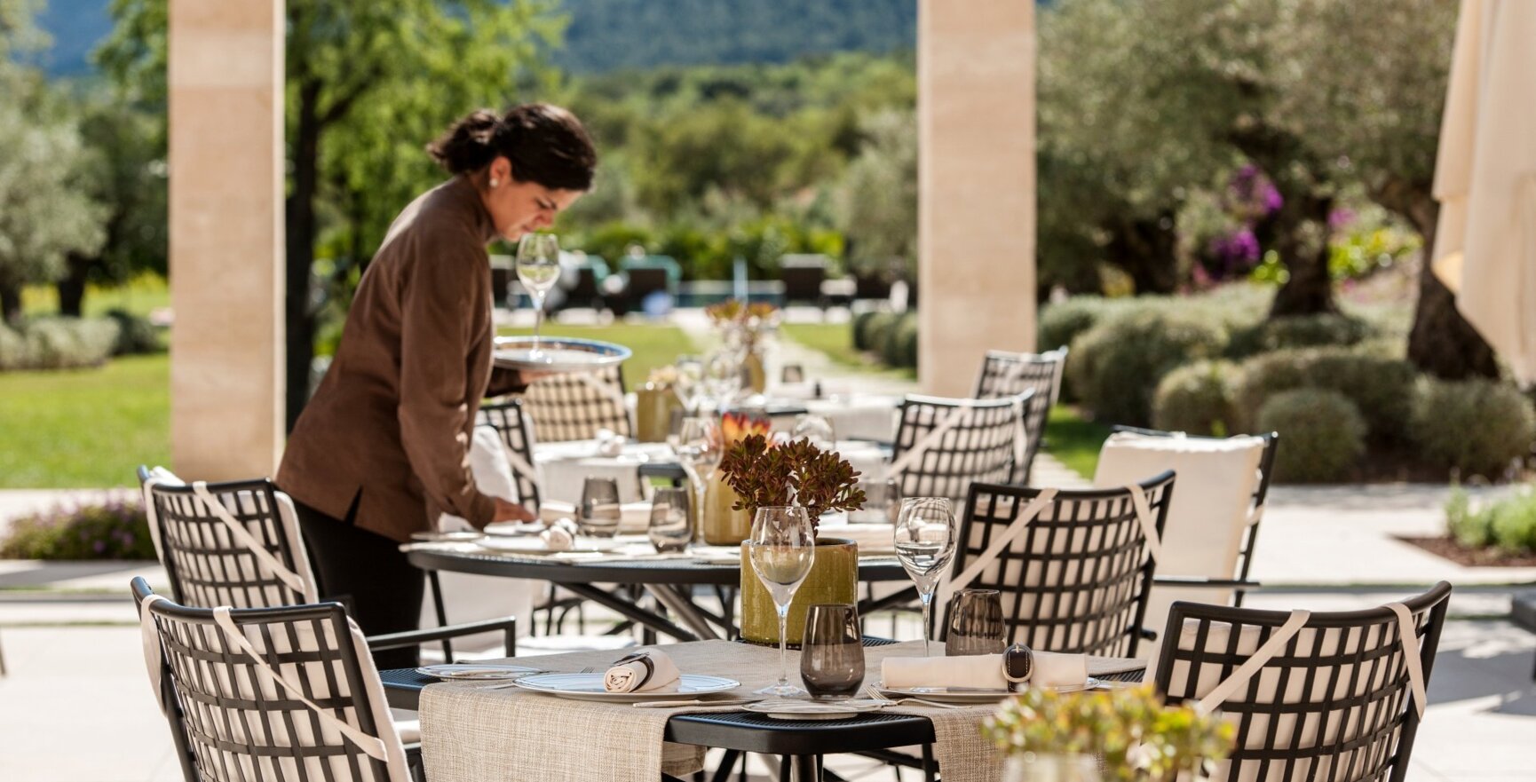 Eine Frau deckt elegant dekorierte runde Tische auf einer sonnigen Hotelterrasse