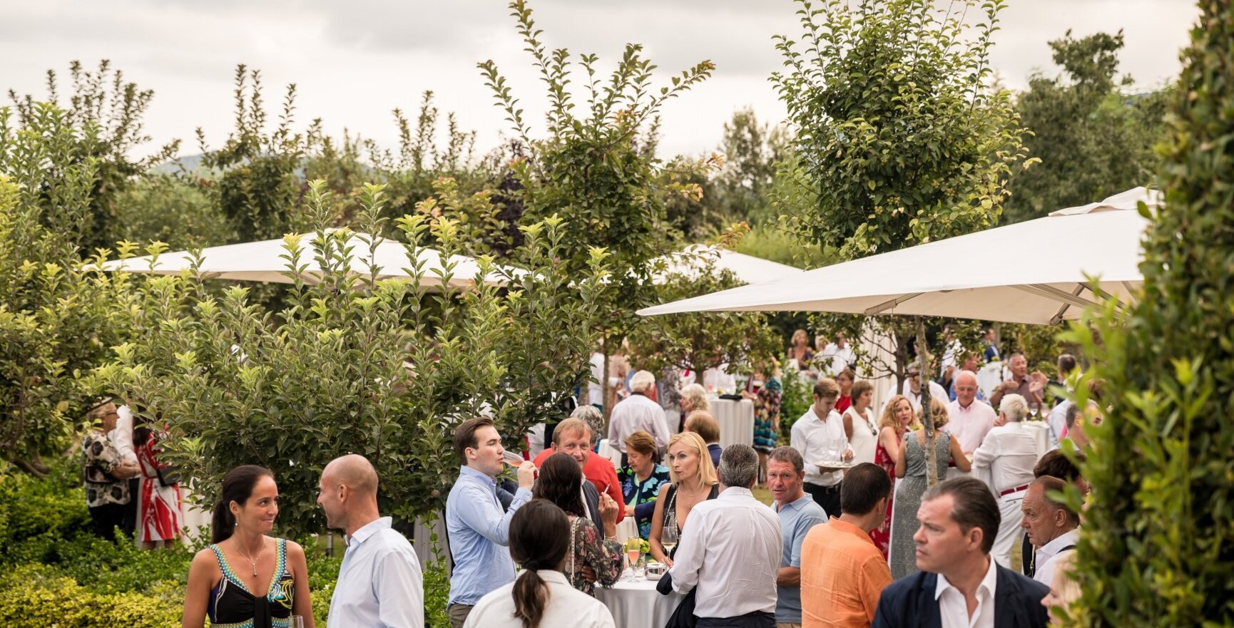 Die Hochzeitsgäste stehen in einem Garten um runde Tische herum und unterhalten sich miteinander