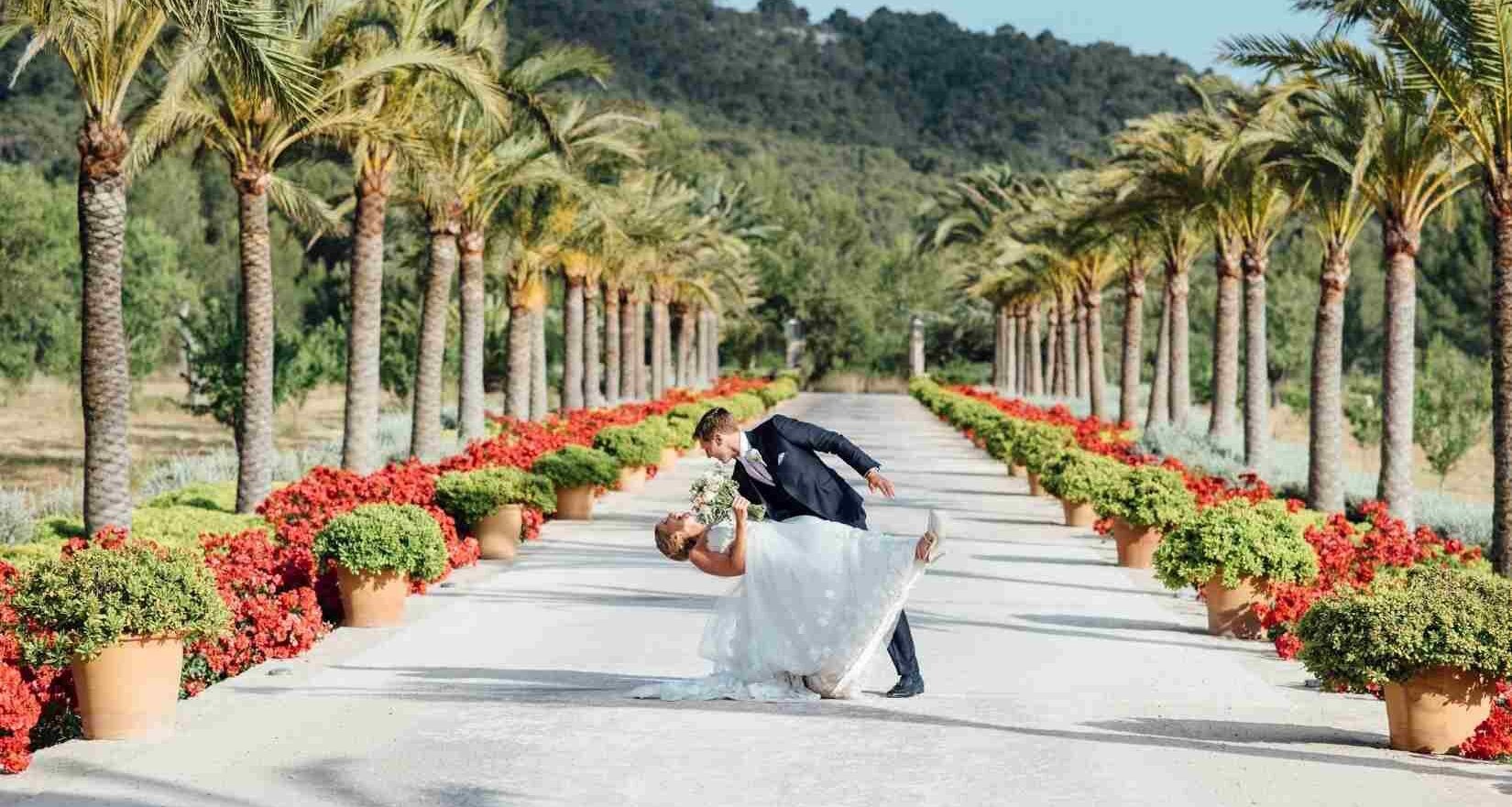 Novia y novio bailando en un camino flanqueado por palmeras, macetas de terracota y flores rojas en una boda al aire libre y soleada
